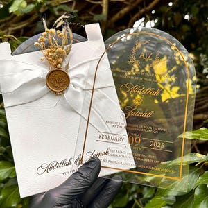 Puede incluir: Invitaci&oacute;n de boda con letras doradas sobre una tarjeta de acr&iacute;lico transparente y una tarjeta blanca con una cinta y un sello de cera. La invitaci&oacute;n incluye los nombres Abdillah & Zainab, la fecha, la hora y el lugar.