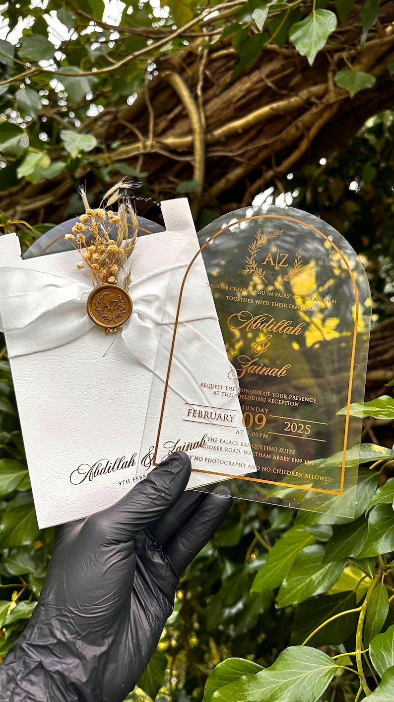 Puede incluir: Invitaci&oacute;n de boda con letras doradas sobre una tarjeta de acr&iacute;lico transparente y una tarjeta blanca con una cinta y un sello de cera. La invitaci&oacute;n incluye los nombres Abdillah & Zainab, la fecha, la hora y el lugar.