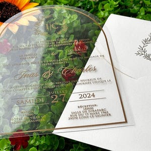 May include: Clear acrylic wedding invitation with gold lettering and a matching envelope. The invitation features the names "Inas & Charles", the date "July 27, 2024", and event details. The envelope has a floral design and the initials "I/C".