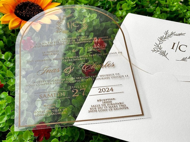 May include: Clear acrylic wedding invitation with gold lettering and a matching envelope. The invitation features the names "Inas & Charles", the date "July 27, 2024", and event details. The envelope has a floral design and the initials "I/C".