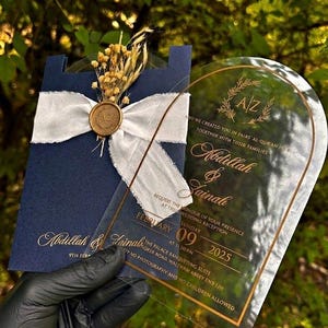 Puede incluir: Invitaci&oacute;n de boda azul marino con escritura dorada, una cinta blanca y un sello de cera dorado. Una invitaci&oacute;n de acr&iacute;lico transparente con texto dorado se sostiene al frente. La invitaci&oacute;n incluye los nombres Abdillah & Zainab y la fecha 09 de febrero de 2025.