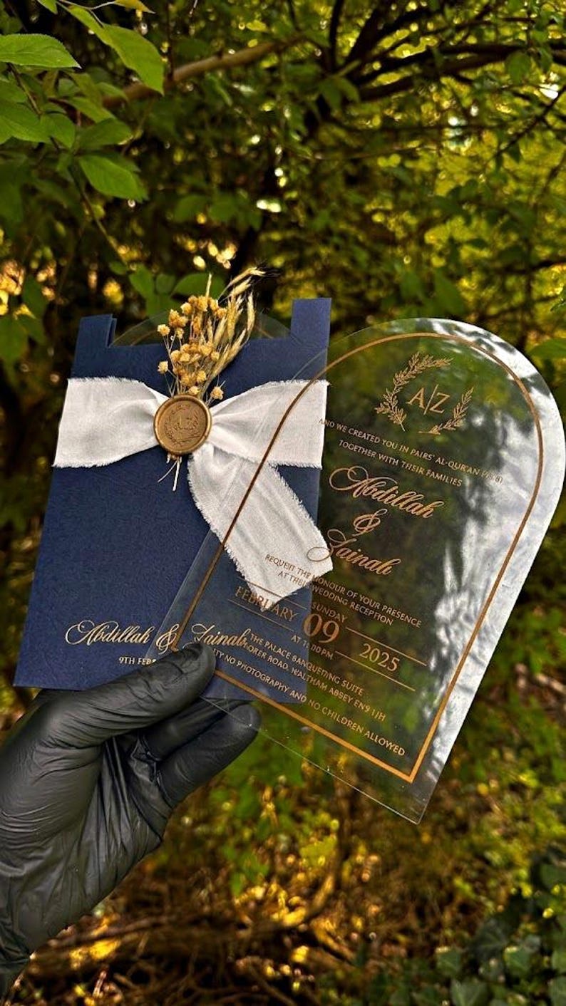 Puede incluir: Invitaci&oacute;n de boda azul marino con escritura dorada, una cinta blanca y un sello de cera dorado. Una invitaci&oacute;n de acr&iacute;lico transparente con texto dorado se sostiene al frente. La invitaci&oacute;n incluye los nombres Abdillah & Zainab y la fecha 09 de febrero de 2025.