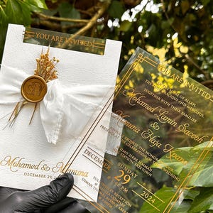 May include: Elegant wedding invitation with gold lettering on a white card, featuring a white ribbon and a gold wax seal. A clear acrylic invitation with gold accents is held next to it. The invitation includes the names Mohamed & Muna, and the date December 29, 2024.