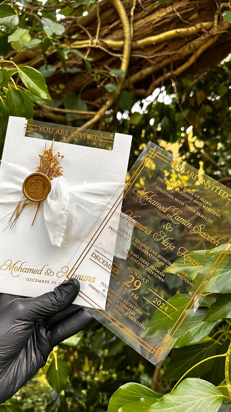 May include: Elegant wedding invitation with gold lettering on a white card, featuring a white ribbon and a gold wax seal. A clear acrylic invitation with gold accents is held next to it. The invitation includes the names Mohamed & Muna, and the date December 29, 2024.