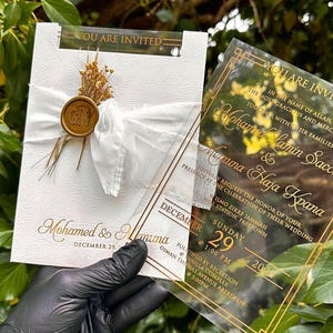 May include: Wedding invitation with gold lettering on a white textured card, tied with a white ribbon and a gold wax seal. A clear acrylic invitation with gold text is held next to it. The text reads "You are invited".