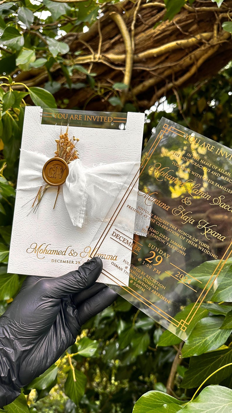May include: Wedding invitation with gold lettering on a white textured card, tied with a white ribbon and a gold wax seal. A clear acrylic invitation with gold text is held next to it. The text reads "You are invited".
