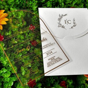May include: Wedding invitation suite featuring a clear acrylic invitation with gold text and a white envelope. The invitation includes the names "Inas & Charles" and the date "2024". A sunflower and greenery are in the background.