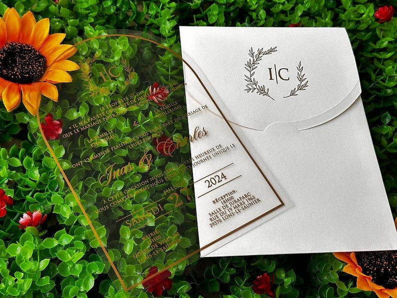 May include: Wedding invitation suite featuring a clear acrylic invitation with gold text and a white envelope. The invitation includes the names "Inas & Charles" and the date "2024". A sunflower and greenery are in the background.