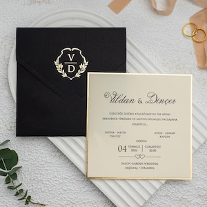 Puede incluir: Un sobre de invitación de boda negro con un emblema dorado y una tarjeta de invitación a juego. La tarjeta tiene un borde dorado y una escritura elegante. Los anillos de boda y la cinta completan la elegante presentación.