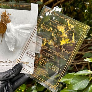 May include: A clear acrylic wedding invitation with gold lettering and a decorative border. The invitation includes the names of the couple, date, and time of the ceremony. A white card with a bow and dried flowers is also visible.
