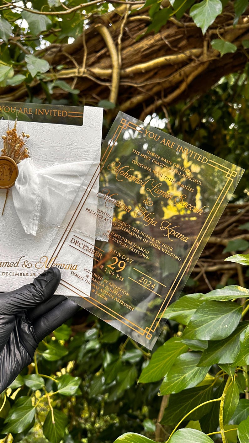 May include: A clear acrylic wedding invitation with gold lettering and a decorative border. The invitation includes the names of the couple, date, and time of the ceremony. A white card with a bow and dried flowers is also visible.