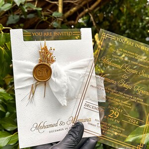 May include: Wedding invitation with gold text on a clear acrylic card and a white card. The white card has a white ribbon, dried flowers, and a gold seal. The text on the cards includes the names Mohamed & Memuna, and the date December 29, 2024.