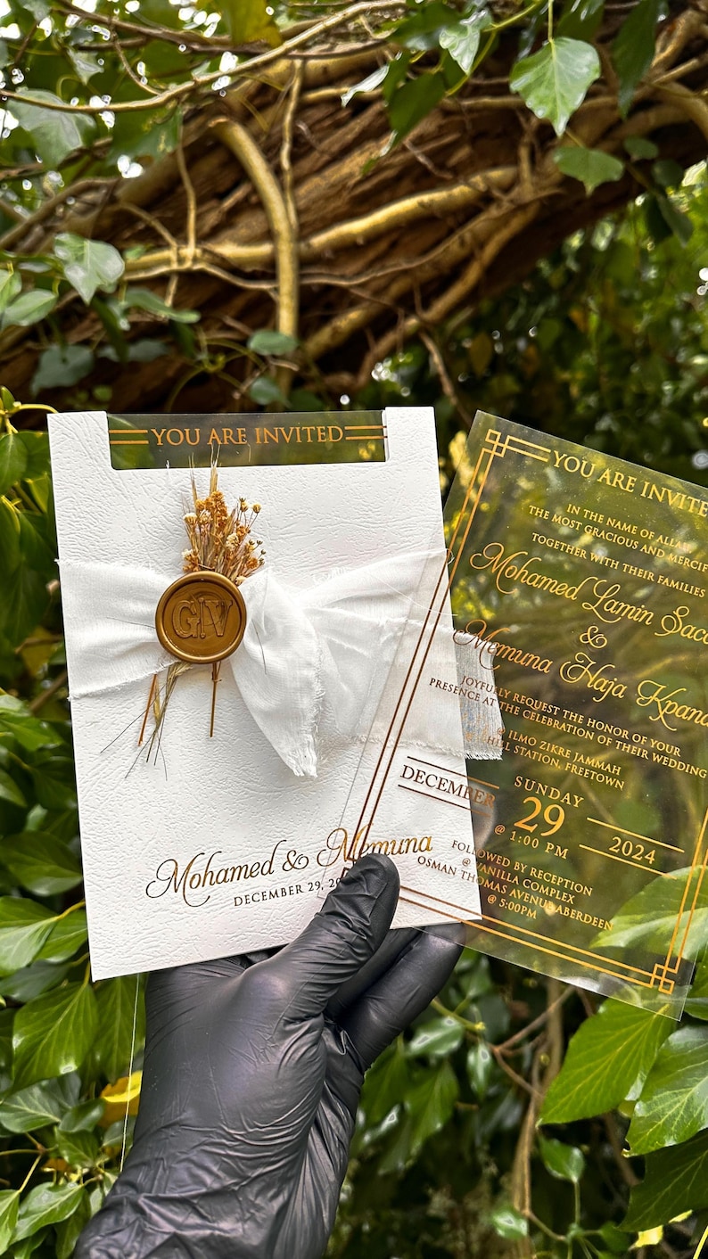 May include: Wedding invitation with gold text on a clear acrylic card and a white card. The white card has a white ribbon, dried flowers, and a gold seal. The text on the cards includes the names Mohamed & Memuna, and the date December 29, 2024.