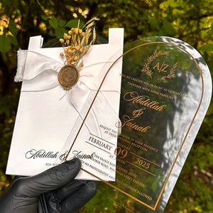 Puede incluir: Elegante invitaci&oacute;n de boda con una tarjeta blanca, una cinta, flores secas y un sello dorado. Una tarjeta de acr&iacute;lico transparente con texto dorado muestra los nombres Abdillah & Zainab, la fecha y los detalles de la recepci&oacute;n.