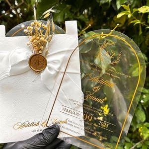 Puede incluir: Invitaci&oacute;n de boda con letras doradas sobre una tarjeta de acr&iacute;lico transparente y una tarjeta blanca. La tarjeta de acr&iacute;lico tiene un borde dorado y los nombres Abdillah & Zainab. La tarjeta blanca est&aacute; decorada con una cinta blanca, flores secas y un sello de cera.