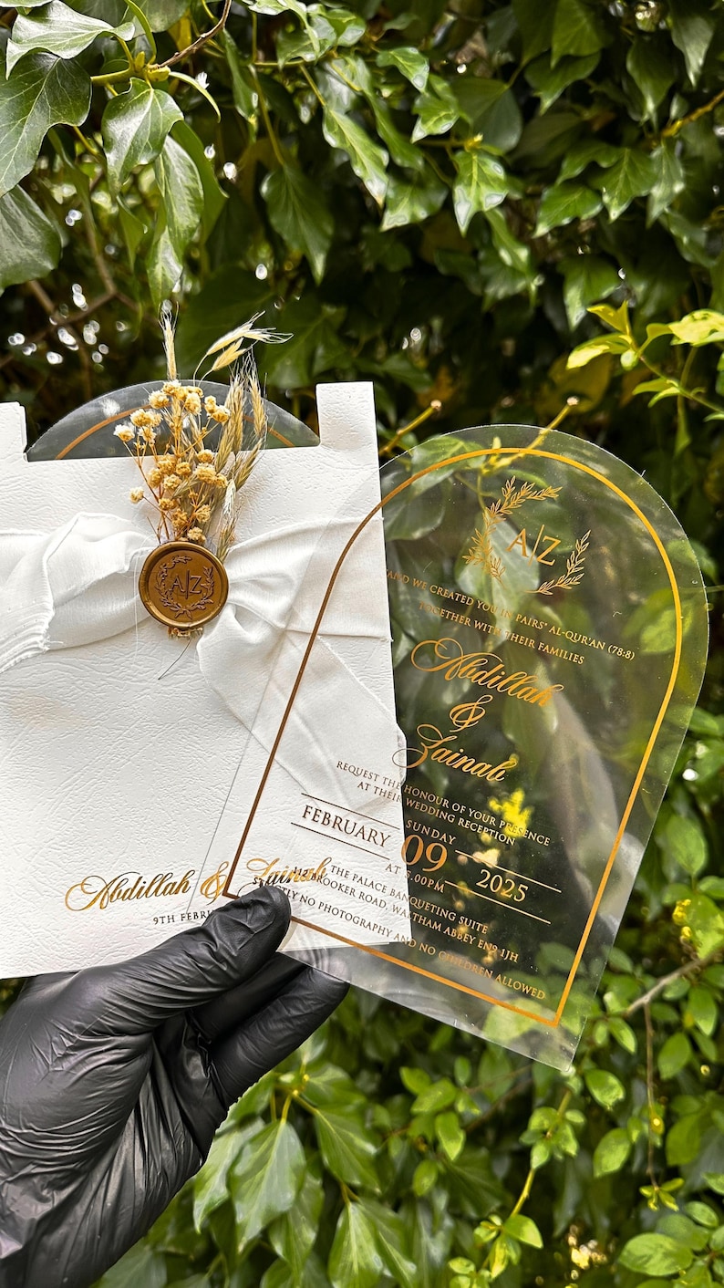 Puede incluir: Invitaci&oacute;n de boda con letras doradas sobre una tarjeta de acr&iacute;lico transparente y una tarjeta blanca. La tarjeta de acr&iacute;lico tiene un borde dorado y los nombres Abdillah & Zainab. La tarjeta blanca est&aacute; decorada con una cinta blanca, flores secas y un sello de cera.