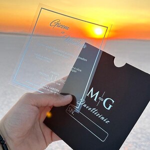 May include: Clear acrylic wedding invitation with silver text, held against a black invitation card. The invitation features names and event details. The background shows a sunset over a white landscape.