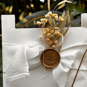 Puede incluir: Tarjeta de invitaci&oacute;n blanca con superficie texturizada, adornada con una cinta blanca y un sello de cera dorado. El sello presenta las iniciales "AZ" y est&aacute; adornado con flores secas y espigas de trigo, creando una est&eacute;tica elegante y natural.
