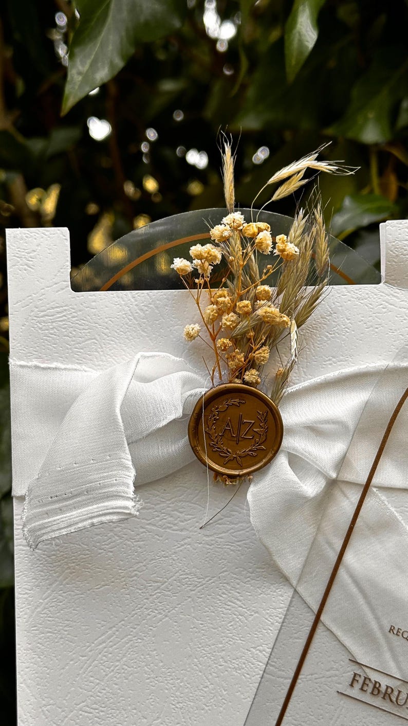Puede incluir: Tarjeta de invitaci&oacute;n blanca con superficie texturizada, adornada con una cinta blanca y un sello de cera dorado. El sello presenta las iniciales "AZ" y est&aacute; adornado con flores secas y espigas de trigo, creando una est&eacute;tica elegante y natural.