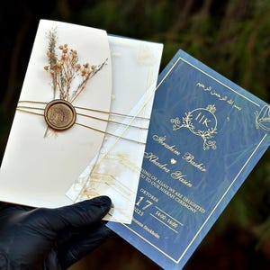Op de afbeelding: Een ivoorkleurige huwelijksuitnodiging met gouden accenten, gedroogde bloemen en een zegel. Een marineblauwe uitnodigingskaart met gouden tekst wordt ernaast gehouden. De tekst bevat namen, datum en tijd.