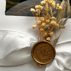 Puede incluir: Una tarjeta blanca con una cinta de raso blanca y un sello de cera dorado. El sello tiene las iniciales "A/Z" y est&aacute; rodeado por una corona de laurel. Flores amarillas secas y espigas de trigo adornan la tarjeta.