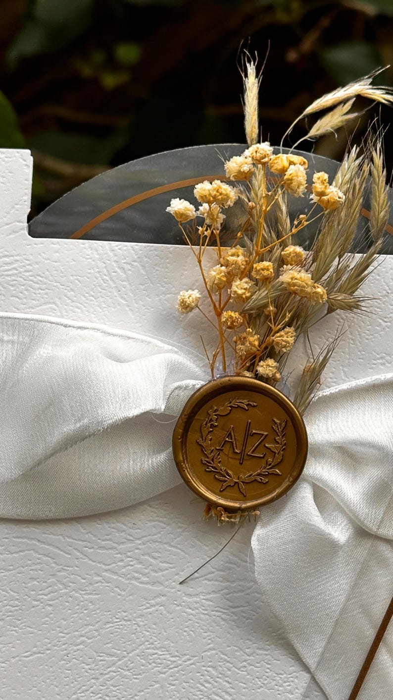 Puede incluir: Una tarjeta blanca con una cinta de raso blanca y un sello de cera dorado. El sello tiene las iniciales "A/Z" y est&aacute; rodeado por una corona de laurel. Flores amarillas secas y espigas de trigo adornan la tarjeta.