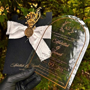 Puede incluir: Una invitaci&oacute;n de boda negra con letras doradas y una superposici&oacute;n de acr&iacute;lico transparente. La invitaci&oacute;n est&aacute; adornada con una cinta blanca, flores secas y un sello dorado. El texto incluye los nombres Abdillah & Zainah y la fecha 09 de febrero de 2025.