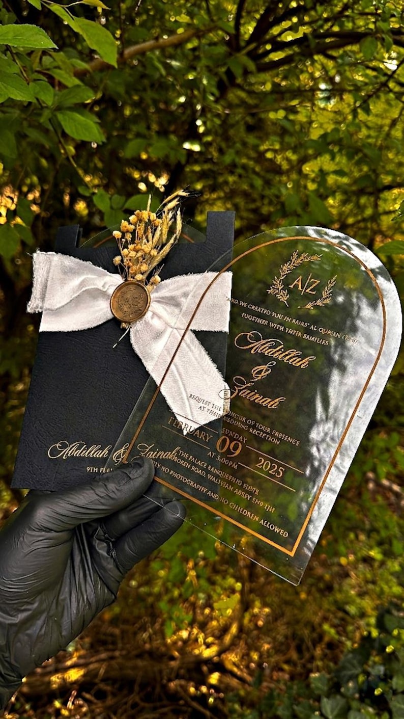 Puede incluir: Una invitaci&oacute;n de boda negra con letras doradas y una superposici&oacute;n de acr&iacute;lico transparente. La invitaci&oacute;n est&aacute; adornada con una cinta blanca, flores secas y un sello dorado. El texto incluye los nombres Abdillah & Zainah y la fecha 09 de febrero de 2025.