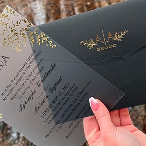 May include: A wedding invitation suite featuring a clear acrylic invitation with gold floral accents and a black envelope. The invitation includes the names of the couple, date, and event details. The envelope has gold initials and the date.