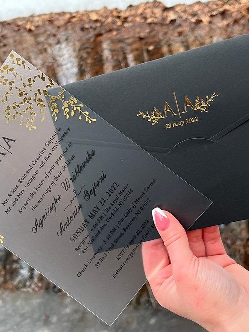 May include: A wedding invitation suite featuring a clear acrylic invitation with gold floral accents and a black envelope. The invitation includes the names of the couple, date, and event details. The envelope has gold initials and the date.