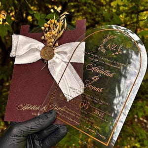 Puede incluir: Una invitaci&oacute;n de boda con una tarjeta burdeos con una cinta blanca y un sello dorado, combinada con una tarjeta de acr&iacute;lico transparente. La invitaci&oacute;n incluye los nombres Abdillah & Zainah, la fecha, hora y lugar.