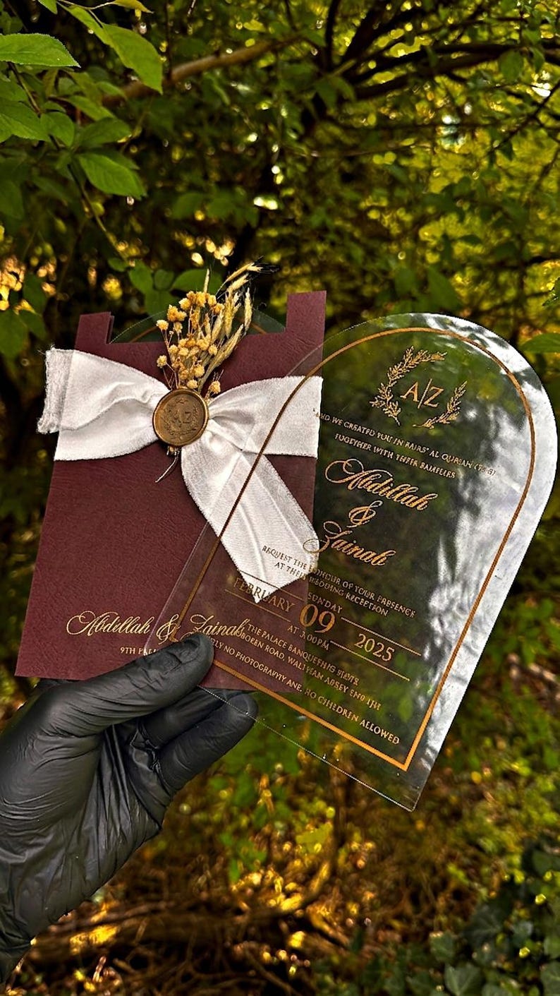 Puede incluir: Una invitaci&oacute;n de boda con una tarjeta burdeos con una cinta blanca y un sello dorado, combinada con una tarjeta de acr&iacute;lico transparente. La invitaci&oacute;n incluye los nombres Abdillah & Zainah, la fecha, hora y lugar.