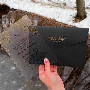 May include: A clear acrylic wedding invitation with gold floral accents and a black envelope. The invitation includes the initials "AA" and the date "22 May 2022". The invitation is held in front of a snowy background.
