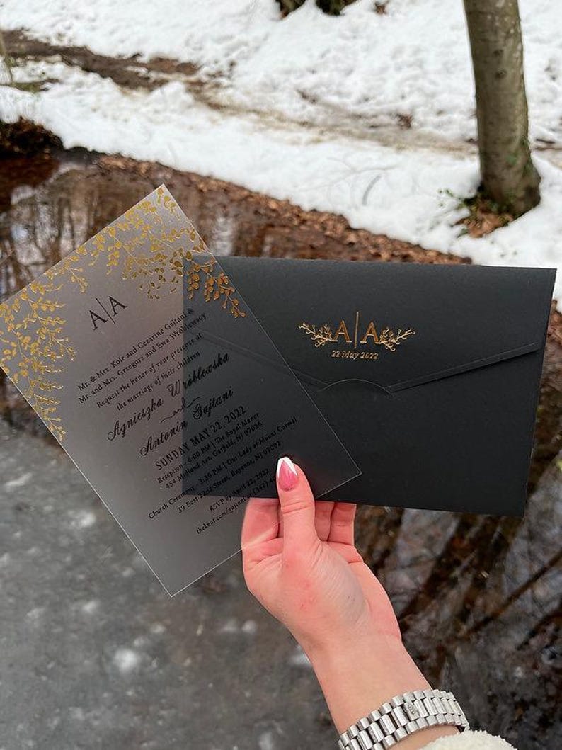 May include: A clear acrylic wedding invitation with gold floral accents and a black envelope. The invitation includes the initials "AA" and the date "22 May 2022". The invitation is held in front of a snowy background.
