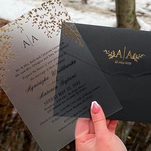 May include: Wedding invitation suite featuring a translucent invitation with gold foil leaf accents and a black envelope. The invitation includes the names Agnieszka and Antonin, the date, and event details. The envelope has the initials "AA" and the date.