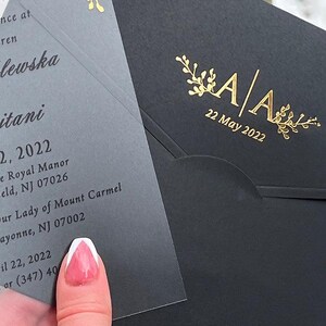 May include: A black wedding invitation envelope with gold foil accents, featuring the initials "A A" and the date "22 May 2022". The invitation card is visible, with text in a delicate font and gold floral details.
