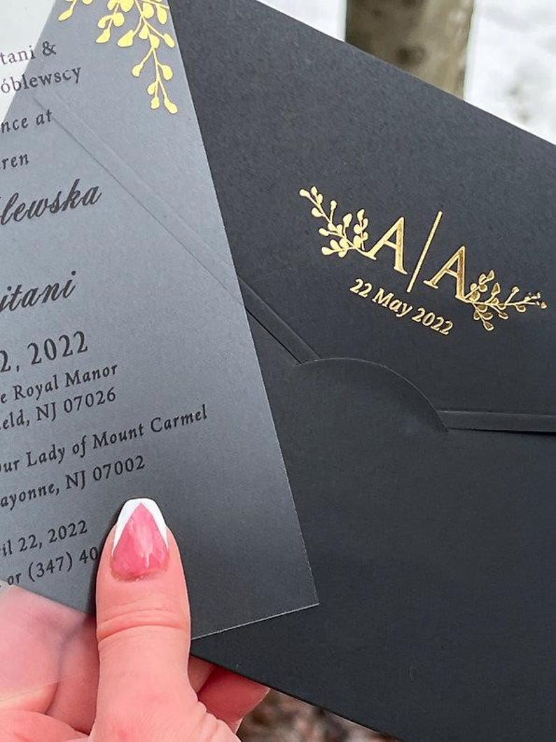 May include: A black wedding invitation envelope with gold foil accents, featuring the initials "A A" and the date "22 May 2022". The invitation card is visible, with text in a delicate font and gold floral details.