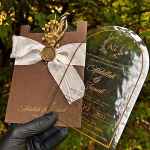 Puede incluir: Una invitaci&oacute;n de boda con una tarjeta marr&oacute;n, una cinta blanca y un sello dorado. La invitaci&oacute;n incluye los nombres Abdullah & Zainab, la fecha, la hora y el lugar. La invitaci&oacute;n se sostiene sobre un fondo verde.