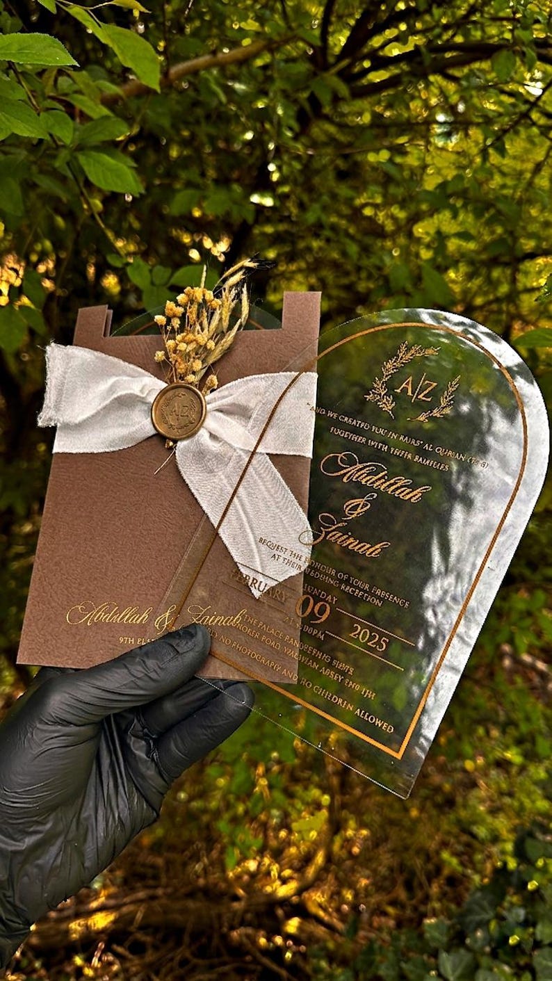 Puede incluir: Una invitaci&oacute;n de boda con una tarjeta marr&oacute;n, una cinta blanca y un sello dorado. La invitaci&oacute;n incluye los nombres Abdullah & Zainab, la fecha, la hora y el lugar. La invitaci&oacute;n se sostiene sobre un fondo verde.