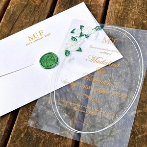 Könnte beinhalten: Hochzeitseinladungs-Set mit einem weißen Umschlag mit grünem Wachssiegel, einer Karte mit floralen Akzenten und einer transparenten Acryl-Einladung. Die Einladung enthält goldfarbene Schrift mit den Namen Madalina und Florentin sowie das Datum 20. August 2023.