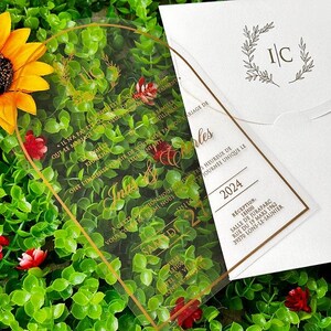 May include: A clear acrylic wedding invitation with gold lettering and a matching white envelope. The invitation features the names "In&egrave;s & Charles" and the date "2024". The background includes greenery and a sunflower.