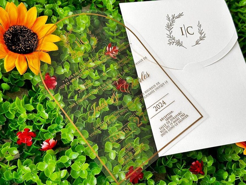 May include: A clear acrylic wedding invitation with gold lettering and a matching white envelope. The invitation features the names "In&egrave;s & Charles" and the date "2024". The background includes greenery and a sunflower.