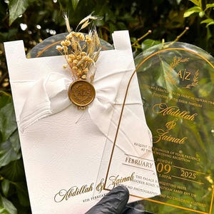 Puede incluir: Elegante invitaci&oacute;n de boda con una tarjeta blanca, lazo, flores secas y un sello dorado. Una invitaci&oacute;n de acr&iacute;lico transparente con texto dorado muestra los nombres Abdillah & Zainab, la fecha y los detalles de la recepci&oacute;n.