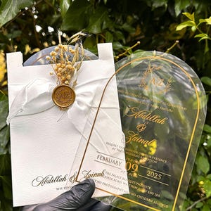 Puede incluir: Elegante invitaci&oacute;n de boda con letras doradas sobre una tarjeta de acr&iacute;lico transparente y una tarjeta blanca con una cinta y flores secas. La invitaci&oacute;n incluye los nombres Abdillah & Zainal y la fecha 9 de febrero de 2025.
