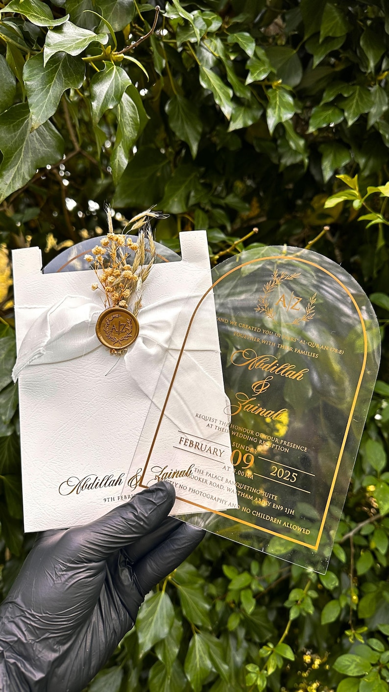 Puede incluir: Elegante invitaci&oacute;n de boda con letras doradas sobre una tarjeta de acr&iacute;lico transparente y una tarjeta blanca con una cinta y flores secas. La invitaci&oacute;n incluye los nombres Abdillah & Zainal y la fecha 9 de febrero de 2025.