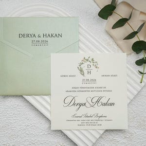 Könnte beinhalten: Hochzeitseinladungs-Set mit hellgrünem Umschlag und cremefarbener Karte, beide mit eleganter Schrift. Die Einladung enthält die Namen "DERYA & HAKAN" und das Datum "27.08.2026". Goldene Schere und dekorative Goldblätter vervollständigen das Arrangement.
