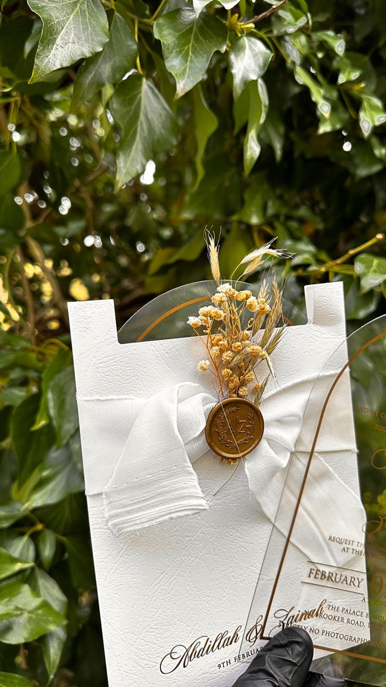 Puede incluir: Una tarjeta de invitaci&oacute;n blanca con una superficie texturizada, adornada con una cinta blanca, flores secas y un sello de cera. La invitaci&oacute;n incluye texto dorado, con una superposici&oacute;n de acr&iacute;lico transparente. El fondo es follaje verde borroso.