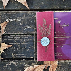 Puede incluir: Invitaciones de boda burdeos y moradas con los nombres "Angélique & Jude" en escritura elegante. Las invitaciones están adornadas con detalles dorados y selladas con un sello de cera azul claro. La fecha "29-12-23" es visible.