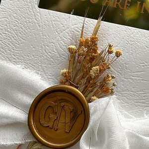 May include: A white textured card with the text "YOU ARE IN" is adorned with a gold wax seal and dried flowers. The seal features the initials "GV". A white ribbon is tied around the card.