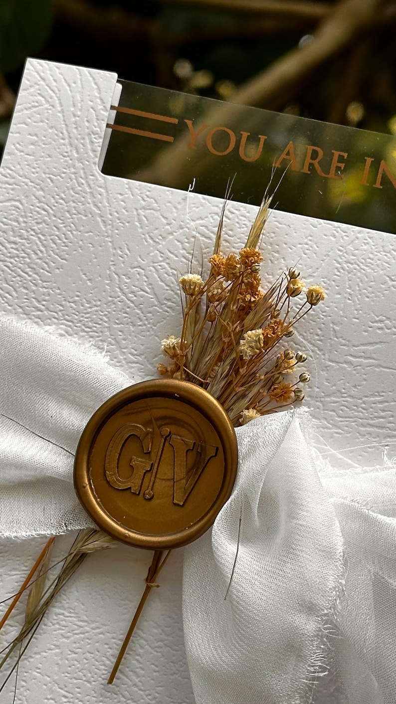 May include: A white textured card with the text "YOU ARE IN" is adorned with a gold wax seal and dried flowers. The seal features the initials "GV". A white ribbon is tied around the card.